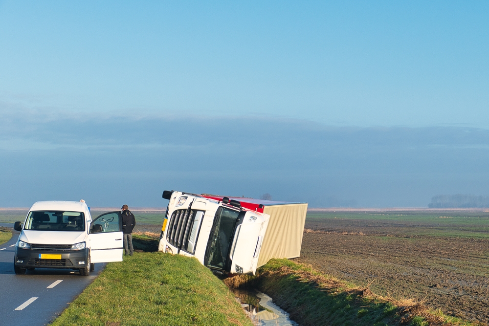 Overturned truck in a ditch in a vast agricultural landscape while the driver is waiting for help.