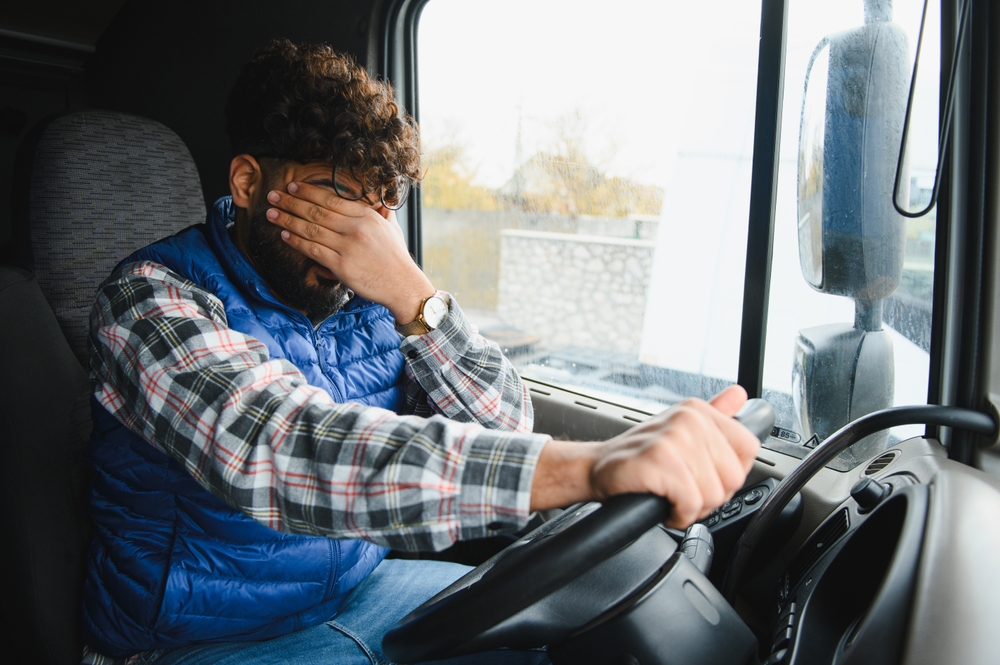 Young male truck driver dealing with exhaustion, stress, and sleep deprivation, rubbing his eyes while on a long journey